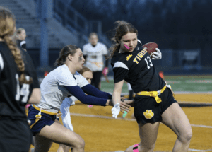 High school girls' flag football game, player in black uniform dodges a defender while carrying the ball. Action shot.