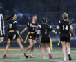 Four female flag football players celebrate during a game, wearing black uniforms with yellow flags on a sports field at dusk.