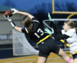 Female athletes competing in a flag football game, one reaching for a catch while being defended by another player.