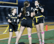 Three girls in black football uniforms celebrating in a night game, showing teamwork and excitement on the field.