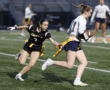 Two female athletes in flag football, one in black and one in white, competing for the ball on the field at dusk.
