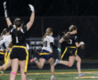 High school girls engage in a flag football game under lights, with one player sprinting toward the end zone.