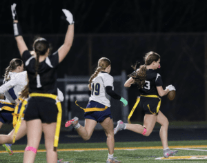 High school girls engage in a flag football game under lights, with one player sprinting toward the end zone.