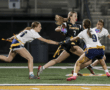 Girls' flag football game action, with a player in black sprinting while being pursued by two defenders in white.