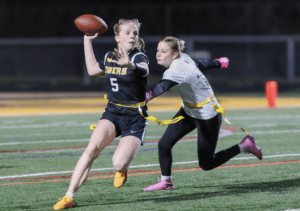 Young female athlete in action, throwing a football while being pursued in a flag football game on a field.
