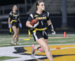 Girl in a Tigers jersey running with a football during a flag football game under stadium lights.