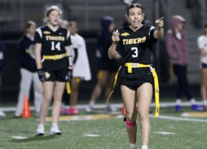 Girls flag football game: a player in a black uniform and yellow belt gestures and communicates strategies on the field.