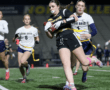 High school girls competing in flag football, with one player advancing the ball while defenders pursue her.