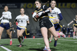 High school girls competing in flag football, with one player advancing the ball while defenders pursue her.