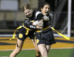 Two young female athletes celebrate a play during a night flag football game, showcasing teamwork and joy.