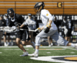 Lacrosse players in action on a field, with one athlete sprinting while the other prepares to catch the ball.