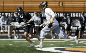 Lacrosse players in action on a field, with one athlete sprinting while the other prepares to catch the ball.