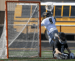 Lacrosse player leaps for a save while another player sits behind the goal, with a school bus in the background.