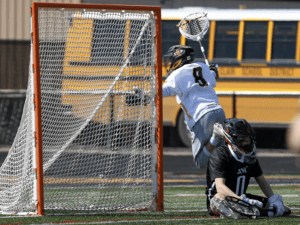 Lacrosse player leaps for a save while another player sits behind the goal, with a school bus in the background.