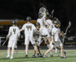 Team celebration after a goal in a lacrosse game, featuring players in white uniforms on a night field.