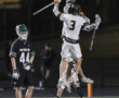 Lacrosse players celebrate a goal, jumping in excitement on the field under stadium lights, with one player in green.