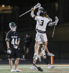 Lacrosse players celebrate a goal, jumping in excitement on the field under stadium lights, with one player in green.