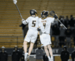 Two lacrosse players in white uniforms celebrate triumphantly after scoring, showcasing teamwork and athleticism.