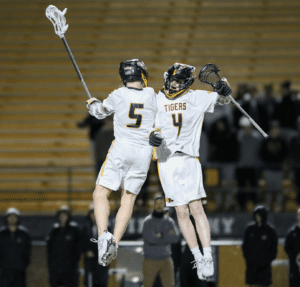 Two lacrosse players in white uniforms celebrate triumphantly after scoring, showcasing teamwork and athleticism.