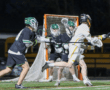 Two lacrosse players compete fiercely as one shoots on goal, with teammates and a net visible in the background.