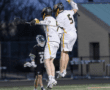 Two lacrosse players in white jerseys celebrate a goal by jumping together, with an opposing player in the background.