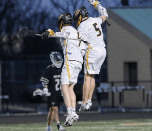 Two lacrosse players in white jerseys celebrate a goal by jumping together, with an opposing player in the background.