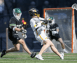 Lacrosse players in action, with one shot on goal while opponents defend during a competitive game at dusk.