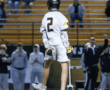 Lacrosse player in a white jersey with the number 2, facing away while fans cheer in the background.
