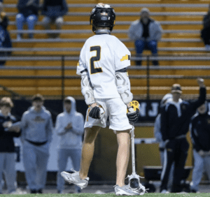 Lacrosse player in a white jersey with the number 2, facing away while fans cheer in the background.