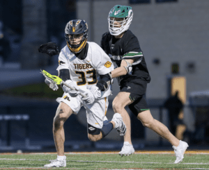 Lacrosse players in action: one in a white Tigers jersey dodging an opponent in black on a turf field.