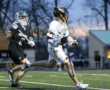 A player in a white lacrosse uniform runs with the ball as an opponent in black tries to defend during a game.