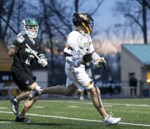 A player in a white lacrosse uniform runs with the ball as an opponent in black tries to defend during a game.