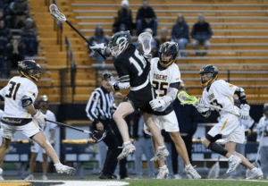 High school lacrosse players compete, showcasing dynamic action on the field during a game. Spectators in the background.