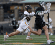 Lacrosse players in action on the field; one player competes vigorously while the other attempts to evade.