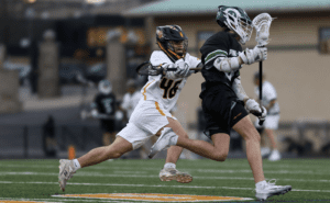 Lacrosse players in action on the field; one player competes vigorously while the other attempts to evade.