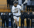 Lacrosse player in action, wearing a Tigers jersey, running on the field with teammates and spectators in the background.