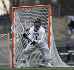 Lacrosse goalie in action, wearing a Tigers uniform, ready to defend the goal on an outdoor field.