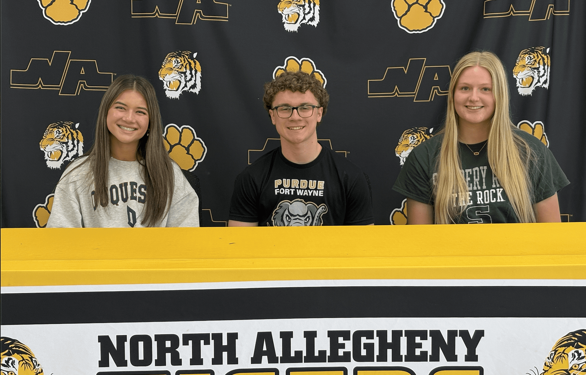 Three students smiling at a signing event, showcasing college commitments at North Allegheny High School.