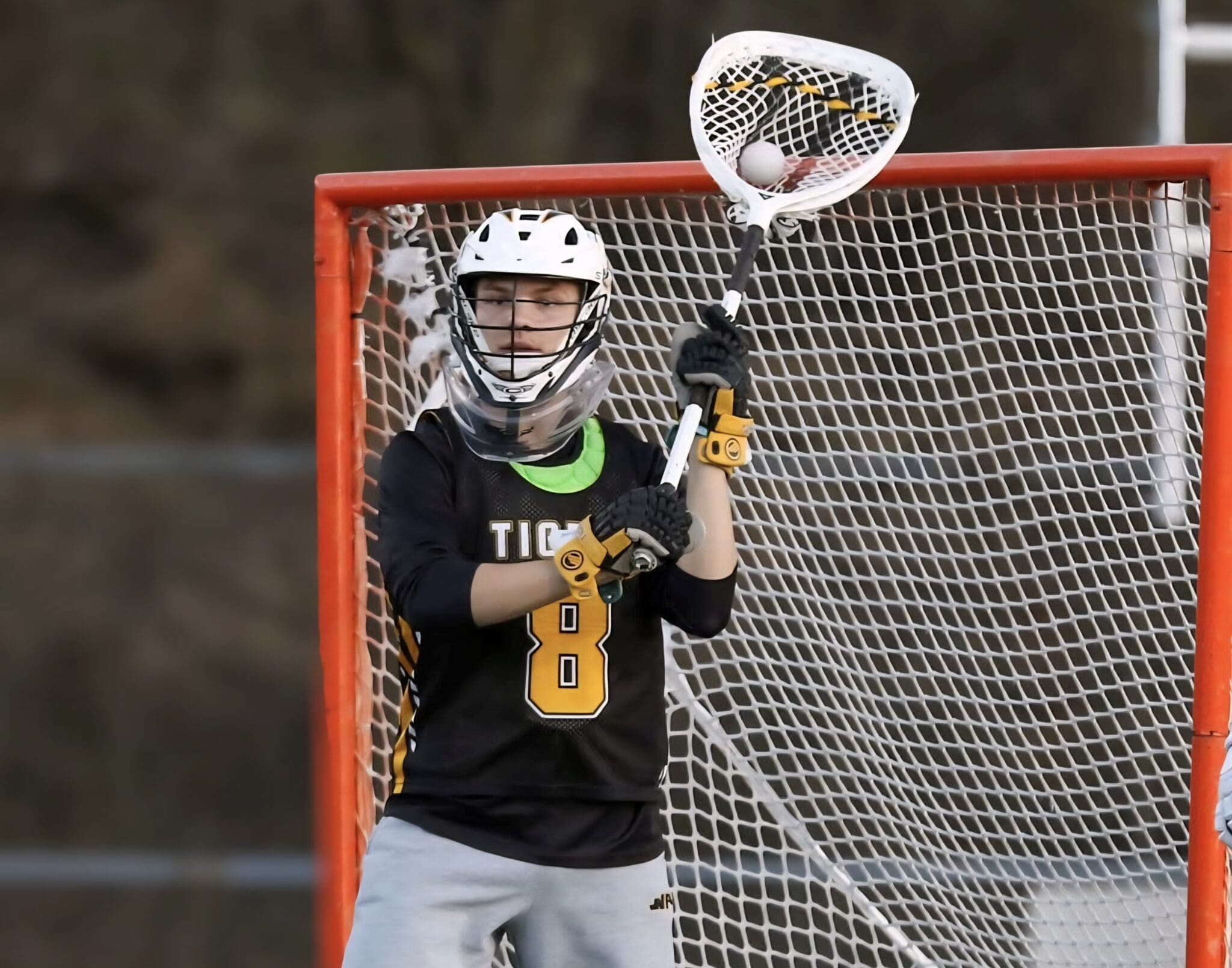 Lacrosse goalie in action, wearing a mask and gloves, ready to block a shot in front of the goal.