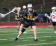 Lacrosse player in yellow and black uniform prepares to pass the ball during an intense game on a turf field.