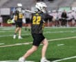 Lacrosse player in black jersey number 22 throws the ball on a field during a game, with teammates in the background.