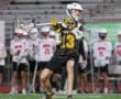 Lacrosse player in action on the field, wearing jersey number 13, as teammates watch from the sidelines.