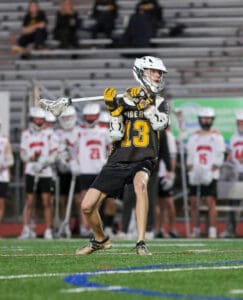 Lacrosse player in action on the field, wearing jersey number 13, as teammates watch from the sidelines.