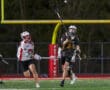 Lacrosse players compete under the lights, one passing the ball while the other attempts to defend.