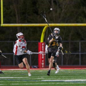 Lacrosse players compete under the lights, one passing the ball while the other attempts to defend.