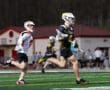 High school lacrosse players sprint during an intense game on a turf field under stadium lights.
