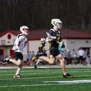 High school lacrosse players sprint during an intense game on a turf field under stadium lights.