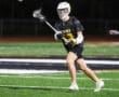 Lacrosse player in action, passing a ball on a night field, wearing a black team jersey with yellow accents.