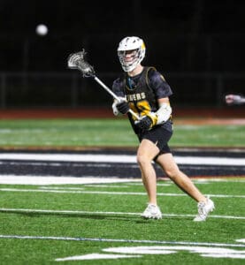 Lacrosse player in action, passing a ball on a night field, wearing a black team jersey with yellow accents.
