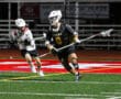 Lacrosse players in action under stadium lights; one in black uniform dodges an opponent in white during a game.
